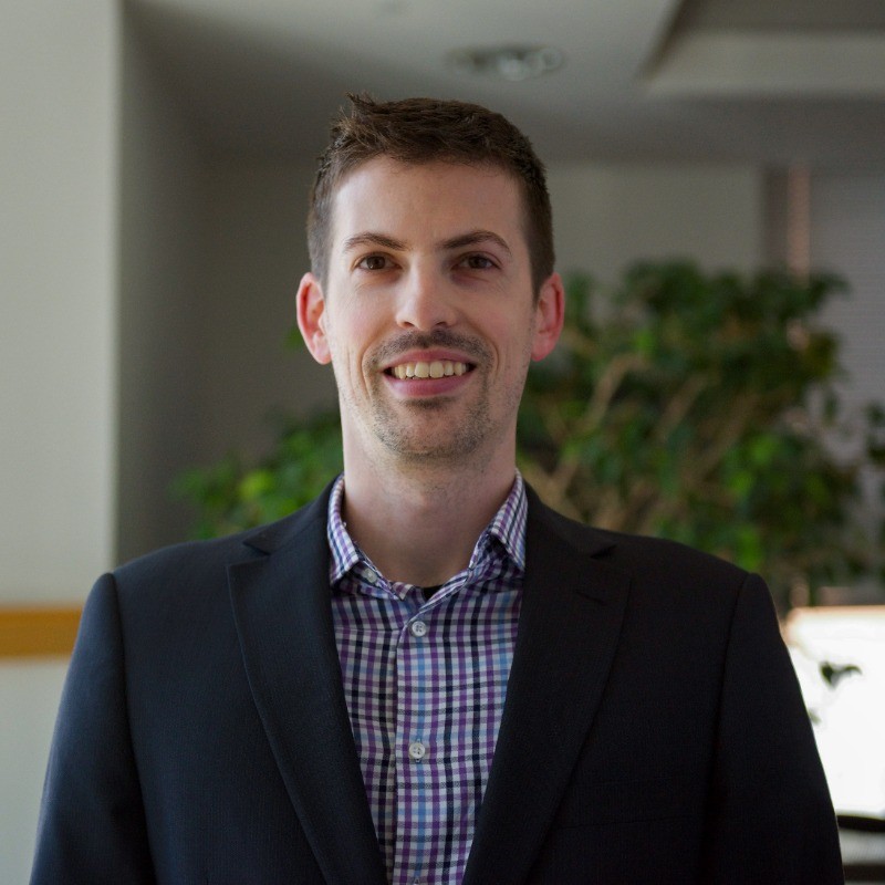 A portrait of Dr. Nic Choquette-Levy, an Assistant Professor of Climate Risk & Decision-Making at Penn State University, smiling while wearing a blazer and checkered shirt, with greenery in the background.